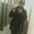 A person in dark clothing and a cap poses with hands near the chest against a blurred background of vertical structures.