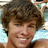 Pixelated close-up of a person with curly brown hair, light skin tone, and visible facial features including eyes, nose, mouth, and chin.