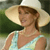 A person wearing a wide-brimmed straw hat and a light blue sleeveless top, with brown hair tied back, set against a blurred green background.