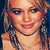 A pixelated image of a woman with long blonde hair in three distinct poses: smiling directly at the viewer, holding a microphone while looking right, and smiling broadly.