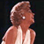 Pixelated image of a person with light brown hair styled in a voluminous updo, wearing a white sleeveless top against a dark background.