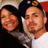 Two people pose together in a selfie-style image with one person wearing a dark cap with a gold emblem and white shirt, the other in dark clothing, against a red and beige background.