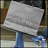 A person holds a gray shopping bag with 'GUCCI' text in white, partially visible blue jeans and dark clothing in the background.