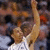 A basketball player in a white jersey with dark patterns raises both arms in a celebratory pose against a blurred crowd background.