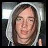 Static image of a person wearing a light gray hooded sweatshirt with visible drawstrings, positioned against an indistinct background featuring blurred architectural elements and lighting fixtures.