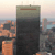 A static pixelated image shows a tall dark building with a brown horizontal stripe near the top, surrounded by smaller orange and gray structures against a light sky background.