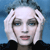 Pixelated portrait of a person with short white hair, blue eyes, and light skin against a blue background, wearing a dark top.