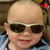 Pixelated close-up of a person with a bald head wearing dark sunglasses and a red hair accessory, smiling slightly against a blurred background.