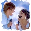 Two people in light blue shirts hold microphones against a bright blue sky background with soft white clouds.