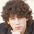 Two side-by-side pixelated images of a person with curly brown hair, wearing dark clothing and holding hands near the chin; one image has a pink heart symbol.