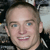 A pixelated close-up of a person's face with light skin, blue eyes, and a slight smile, set against a blurred background with dark and light tones.