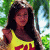 A pixelated image of a person in a yellow top with red accents, standing outdoors near green foliage and a light-colored structure.