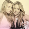 Two women with long blonde hair pose closely together, one in a pink sleeveless top, the other in a black top with a white pendant necklace, against a light background.