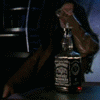 A person with long dark hair drinks from a glass while holding a bottle with a black label featuring white text and a red dot, under blue neon lighting.