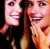 Two women with long hair smile; one covers her mouth. Pink background has text and small purple/black symbols.
