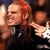 Pixelated static image of a person with long red hair wearing dark clothing, holding an object near their face against a dark background.