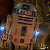 Pixelated static image of a droid-like character with metallic brown and blue coloring, orange sensor light, and glowing blue accents on arms against dark background.