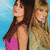 Two women in casual clothing stand side-by-side against a blue background, one with long brown hair and the other with blonde hair.