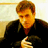 Pixelated static image of a person with light brown hair wearing dark clothing, holding an object in their hands against a gradient background.