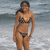 A woman in a black bikini stands on a beach with ocean waves behind her, showing slight variations