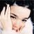 Pixelated close-up of a person with light skin and dark hair, wearing a white top, holding a hand near their head against a dark background.