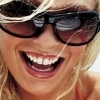Close-up of a person with blonde hair wearing dark sunglasses, showing a wide open mouth with visible teeth and a red lip color.