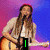 A person with long brown hair plays an acoustic guitar on a stage, wearing a light-colored cardigan over a white top against a purple and blue gradient background.