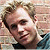 Pixelated static image of a person's face with light brown hair and visible facial features including eyes, nose, mouth, and ear, wearing dark clothing against a blurred background.