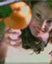 A woman with long dark hair holds an orange fruit near her face, smiling slightly while wearing a light-colored top.