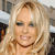Pixelated close-up of a person with long blonde hair wearing a dark choker necklace, smiling with visible teeth against a blurred background.