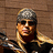 Pixelated static image of a person with long blonde hair wearing dark sunglasses and a gray beanie, set against a brown wooden background.