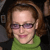 Pixelated close-up of a person with brown hair wearing purple glasses and a green turtleneck sweater against a dark background.