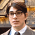 Pixelated portrait of a person with dark hair wearing glasses, a white shirt, and a dark tie against a blurred background with warm tones.