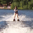 A person stands on water skis in a lake, holding a tow rope, with trees in the background.