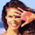 Pixelated static image of a woman with brown hair wearing a black top, smiling and touching her face against a light blue background.