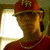 Pixelated image of a person wearing red cap and shirt, gold chain necklace, dark sunglasses, standing near bright light source with greenish background.
