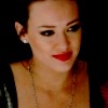 A close-up of a person with dark hair pulled back, wearing red lipstick, a silver earring, and a gold chain necklace against a dark background.