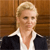 Pixelated image of a person with short blonde hair wearing a white collared shirt and dark jacket against a brown wooden background.
