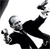 Pixelated grayscale image of a figure in a black suit performing a dynamic pose with arms raised and legs spread against a white background.