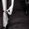 Black-and-white close-up of a person's lower legs and feet wearing high-heeled shoes, with one hand adjusting a bracelet on the ankle.