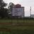Pixelated outdoor scene showing a large sign structure with dark top and light base, surrounded by trees and grass under overcast sky.