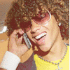 A person with curly brown hair wears pink sunglasses, a yellow shirt, and a purple jacket while holding a silver phone to their ear.