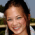 A pixelated close-up of a person with dark hair tied back, smiling broadly with eyes partially closed, wearing a light-colored top against a blurred outdoor background.
