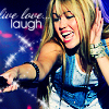 A woman with long blonde hair and bangs smiles broadly while holding a microphone, wearing a blue sleeveless top and white shorts against a dark background with 'live laugh' text.