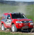 A red pickup truck with a white front bumper and silver accents is positioned on a grassy field under a cloudy sky.