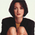 Pixelated image of a person with short dark hair wearing a black jacket over a light brown shirt, hands clasped in front, against a plain light background.