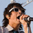 Pixelated image of a person with long dark hair wearing sunglasses and holding a microphone, next to text reading "Joe Jonas (33)" on a black background.