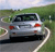 A silver sedan with red taillights drives on a winding road through green fields under a light blue sky.