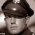 Static grayscale icon of a person wearing a military-style cap with a small emblem, cropped to show head and shoulders against a light background.