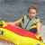 A person in a gray and yellow life vest holds a red and yellow inflatable raft on water, with the right arm extended forward.